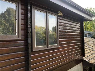 Close-up view of a house with dark wood siding and two large windows, reflecting the surrounding greenery.