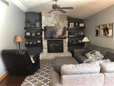 A cozy living room with a stone fireplace, mounted TV, gray sectional sofa, black leather recliner, floating shelves with decor, and a patterned rug.