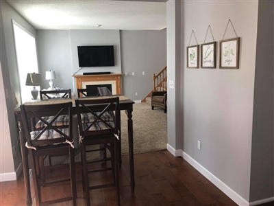 A dining area with a high table and chairs, adjacent to a living room with a fireplace and wall-mounted TV, and three framed pictures on a gray wall.