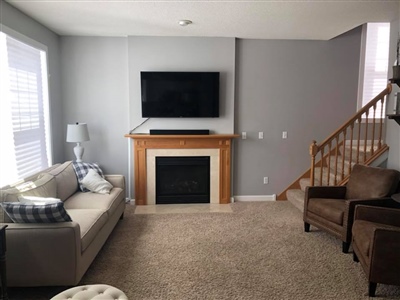 A cozy living room with a beige sofa, a brown armchair, a fireplace with a wooden mantle, a wall-mounted TV, and carpeted flooring.