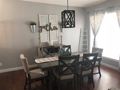 A dining room with a dark wooden table, six chairs, a modern chandelier, gray walls, and a large window with sheer curtains.