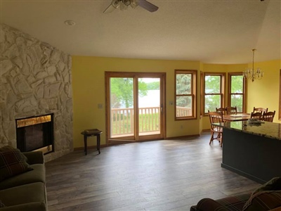 An open living area with yellow walls, a stone fireplace, large windows and glass doors leading to a deck, and a kitchen island with seating.