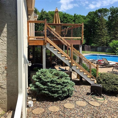 Outdoor area with a wooden deck and staircase, leading to a patio with a swimming pool, surrounded by greenery and landscaping.