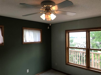 A room with green walls, a ceiling fan with lights, two windows, and a sliding glass door leading to an outdoor deck.