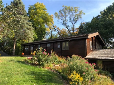 A single-story house with dark wood siding, surrounded by a lush garden and green lawn, with trees and a clear blue sky in the background.