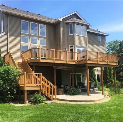 Two-story house with a large wooden deck and staircase, a patio underneath, and a well-maintained lawn, surrounded by greenery.