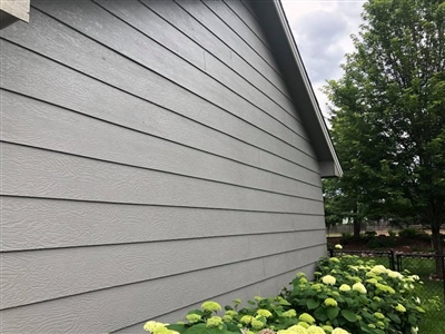 Side of a gray house with horizontal siding, surrounded by green bushes and trees.