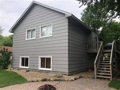 A gray house with horizontal siding, multiple windows, a staircase leading to a deck, and a landscaped yard with a fire pit.