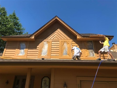 Two workers painting the upper exterior of a wooden house with tan siding, on a sunny day, with trees in the background.