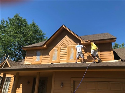 Two workers painting the upper exterior of a wooden house with orange-brown siding on a sunny day, with trees in the background.