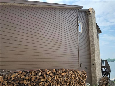 Side of a brown house with horizontal siding, a stack of firewood against the wall, and a stone chimney, with a view of the lake in the background.