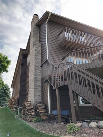 Side view of a brown house with a stone chimney, an external staircase leading to a balcony, and stacks of firewood along the wall.