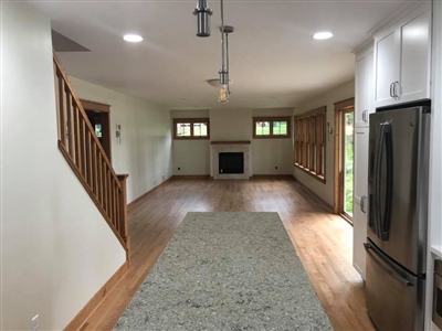 Open living area with wooden flooring, a staircase with wooden railings, a fireplace, multiple windows, and a kitchen with a stainless steel refrigerator.