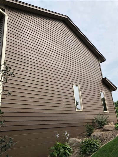 Side of a brown house with horizontal siding, small windows, and a landscaped garden with plants and flowers.