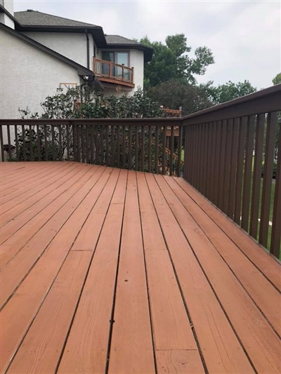 A wooden deck with reddish-brown planks and a brown railing, overlooking a garden and neighboring house.