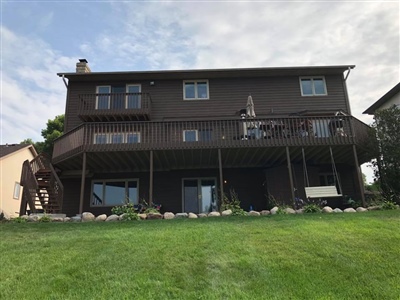 A two-story house with dark brown siding, a large elevated deck, and a covered patio area below, surrounded by a well-maintained lawn.