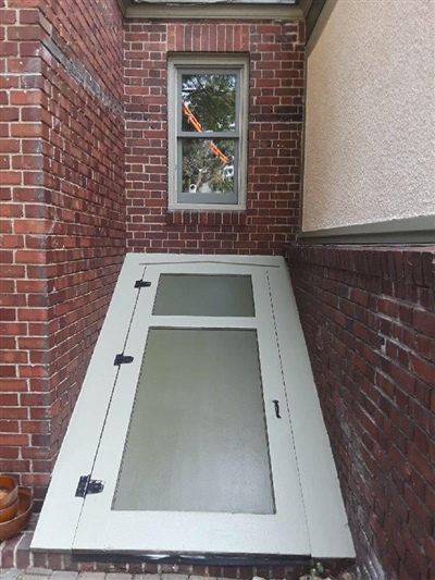 A brick exterior wall with a green cellar door and a window above it.