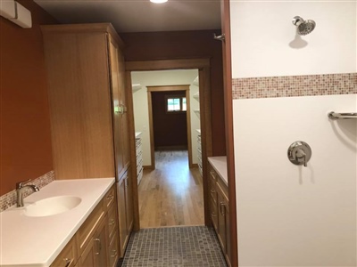 A bathroom with wooden cabinetry, a white countertop, a walk-in shower, and a view through to a hallway with wooden flooring.