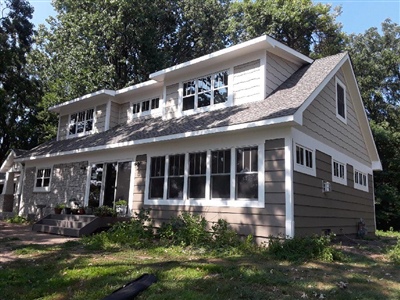 A two-story house with a mix of beige and brown siding, large windows, and a sloped roof, surrounded by trees and greenery.