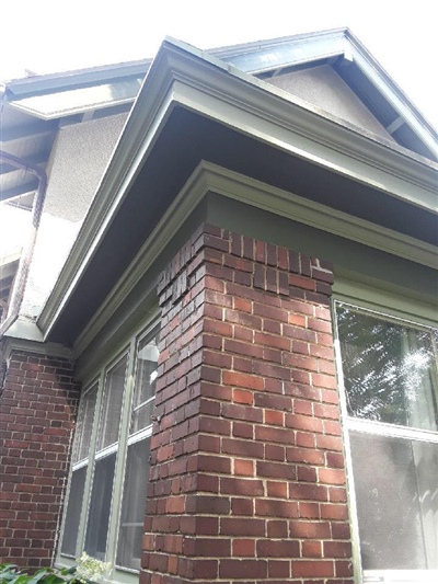 A brick exterior corner of a house with green trim and large windows, showing the overhang of the roof and part of the upper story.