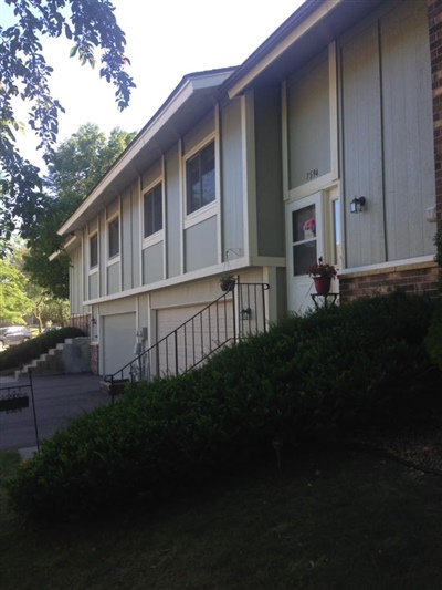 A single-story house with light-colored siding, large windows, and a staircase leading to the front door, surrounded by greenery.