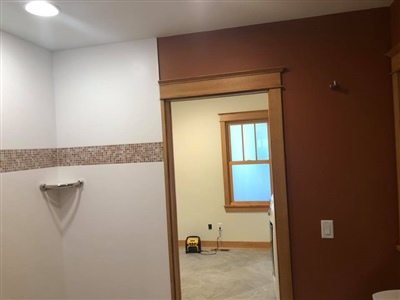 A bathroom with white and brown walls, a tiled section with a decorative border, and a view into an adjacent room with beige carpet and a window.