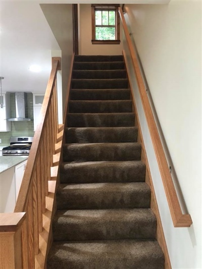 A carpeted staircase with wooden railings leading up to a small window, with a view of the kitchen area to the side.