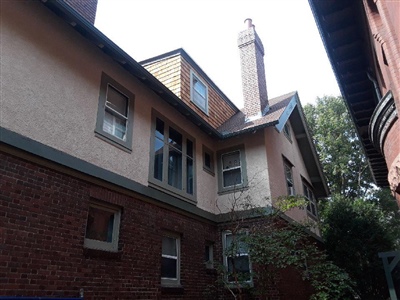 A multi-story house with a combination of brick and stucco exterior, featuring multiple windows and a chimney, surrounded by trees and neighboring buildings.