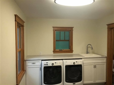 A laundry room with a front-loading washer and dryer, a countertop, a sink, and wooden-trimmed windows.