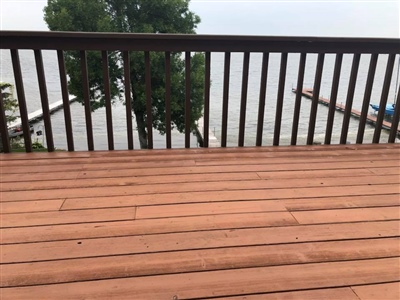 A wooden deck with a brown railing overlooking a body of water with trees and a dock in the background.