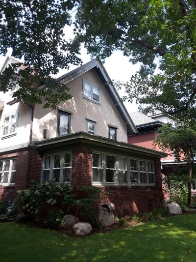 A two-story house with a brick lower level and stucco upper level, surrounded by trees and a well-maintained lawn with shrubs and rocks.