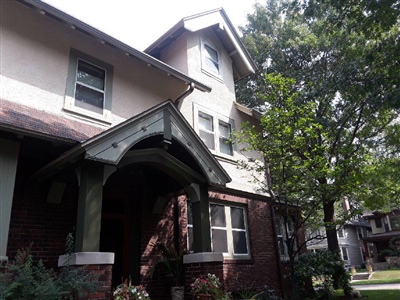 A two-story house with a brick lower level and stucco upper level, featuring a covered entrance with green trim, surrounded by trees and greenery.