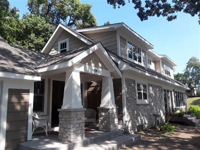 A two-story house with beige siding, stone accents, a covered front porch, and white trim, surrounded by trees and a landscaped yard.