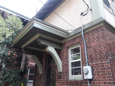 A close-up of a house exterior with a brick lower level and stucco upper level, featuring green trim and an overhang with decorative brackets.