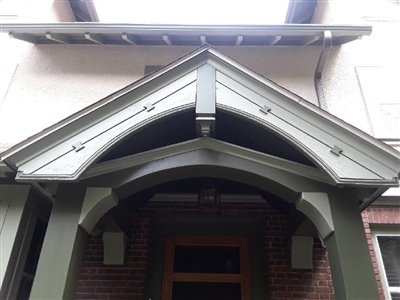 A close-up of a house entrance with a green gabled overhang, decorative trim, and brick lower walls, highlighting the architectural details.