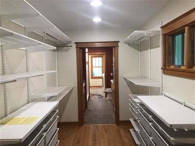 A walk-in closet with white shelving and drawers, wooden trim, hardwood flooring, and recessed lighting, leading to a hallway with carpeted flooring.