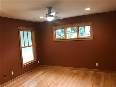 A room with red walls, wooden flooring, recessed lighting, a ceiling fan, and windows with wooden trim.