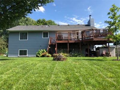 A two-story house with blue siding, a wooden deck, a well-maintained lawn, and trees in the background.