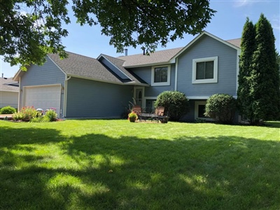 A two-story house with blue siding, a well-maintained lawn, a two-car garage, and trees and shrubs surrounding the property.