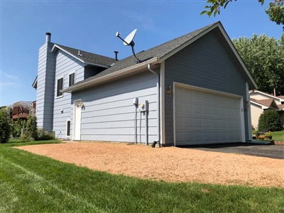 A two-story house with blue siding, a two-car garage, a satellite dish on the roof, and a gravel driveway, surrounded by a well-maintained lawn.