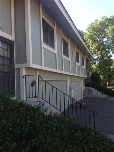 A single-story house with light gray and white siding, large windows, and a staircase leading to the front door, with a driveway and surrounding greenery.