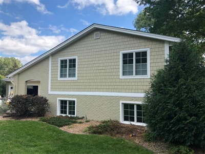 A two-story house with light yellow siding and white trim, multiple windows, and a landscaped yard with shrubs and trees.