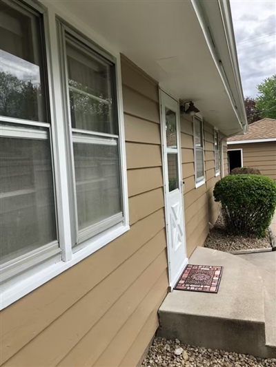 A beige house exterior with horizontal siding, white-framed windows, a white door with a welcome mat, and a small shrub beside a concrete walkway.