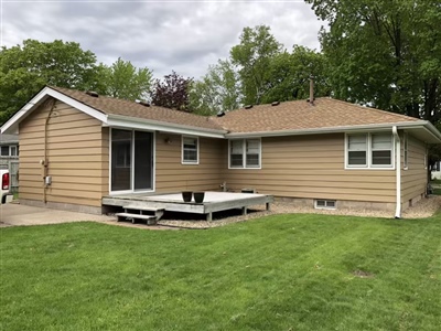 A single-story house with beige siding, a sliding glass door leading to a small wooden deck, and a well-maintained lawn, surrounded by trees.