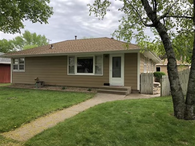 A small beige house with a brown roof, white front door, large window, green lawn, tree on the right, and a concrete path leading to the entrance.