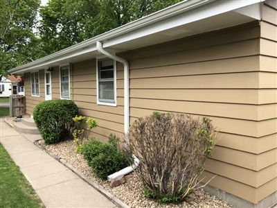 Side view of a beige house with white trim, multiple windows, neatly trimmed bushes, and a concrete walkway beside the house.