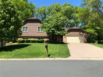 Two-story brown house with a large front lawn, trees, and a concrete driveway leading to a two-car garage.