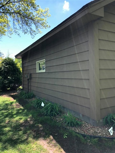 Side view of a brown house with a small window, surrounded by green grass, plants, and trees under a clear sky.