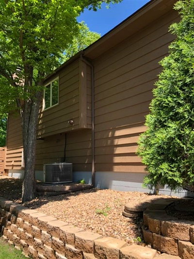 Side view of a brown house with a small window, a tree, a neatly landscaped area with mulch and a retaining wall, and green shrubs.