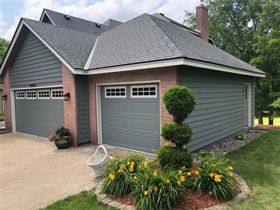 Gray house with brick accents, a two-car garage, manicured landscaping with a topiary, flowers, and a concrete driveway.